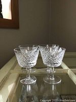 Four clear Waterford crystal glasses with cut pattern, displayed on a reflective glass surface.