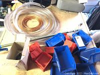 Photo showing assorted red and blue plastic storage bins, coiled plastic and copper tubing, and metal grabber tool on a workbench, with some cardboard box and other items in background