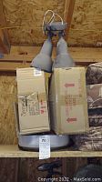 Outdoor dual spotlight with two large metal cone shades, hanging from ceiling above two cardboard boxes.