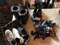 Top-down view showing white Nike shoes, black snowboard boots, multiple pairs of men's shoes inside a woven basket along with suspenders.