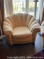 Clamshell-style upholstered armchair in light beige upholstery, shown on hardwood floor in front of window.