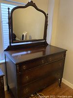 Photo shows a vintage solid wood dresser with an attached tall mirror featuring a decorative carved top. The dresser has five drawers with round metal ring pull handles and turned legs. The wood finish is polished dark brown with some signs of age.