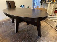 Photo of a large black oval dining room table with two removable leaves, placed on a wooden floor with visible wear. One leg shows veneer damage.