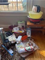 Photo of the entire assortment including red electric wok, glass lid, yellow bowl, popcorn maker box, Slap Chop box, Stanley thermos, egg coolers, glass pitcher, and various kitchen tools on carpet and small bench.