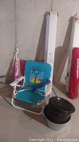 Image showing a turquoise nylon folding beach chair with palm tree design, black enamel lobster pot, and other beach gear in background.