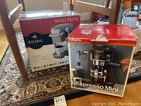 Wide view of both Keurig Mini Plus and Krups Espresso Mini boxed machines on floor under table.