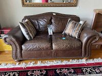 Front view of worn brown leather loveseat with two striped throw pillows and sealed leather cleaner pack on seat cushions. Shows detail of nailhead trim and rolled armrests.