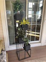 Four-tier black metal plant stand with four faux plants on top in black square pots, placed on porch deck against windowed door.