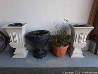 Full view of four planters arranged side by side showing two tall white concrete planters, one black round plastic planter, and one orange plastic planter with plant.