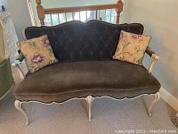 Front view of the dark brown velvet upholstered settee with tufted backrest, cream-colored wooden legs and armrests, and two floral-patterned throw pillows.