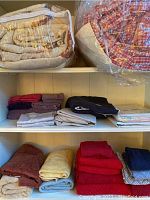 Photo of shelves showing beige comforter and red plaid comforter in plastic bags, folded sheets and towels in various colors.