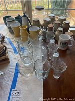 Photo shows a variety of ceramic spice storage canisters with cork lids, yellow and blue ceramic teapots, ceramic and glass mugs, and glassware arranged on a table.
