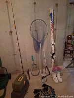 Fishing rods, tennis racquet, fishing net, Salomon skis and boots, lacrosse stick, roller blades seen against basement concrete wall