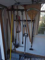 Photo of garden tools hanging in garage including rakes, shovels, hoes, post hole digger, bow saw, and push broom.
