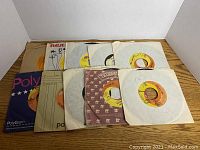 Top down image of 10 vintage 45 RPM vinyl records in assorted paper sleeves laid out on a wooden surface.