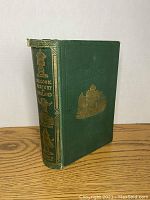 Book standing upright revealing green cloth covering and gold embossed figures and titles on spine. Shows wear and slight detachment of cover from binding.