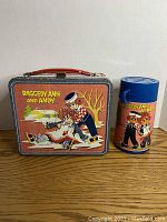 Front view of the Raggedy Ann and Andy lunchbox with colorful character scene, alongside matching thermos including lid and artwork