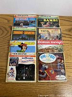 Photo showing eight vintage View-Master reels in original sleeves arranged in two columns on wood surface. Titles visible include Disney's Bambi, Mickey Mouse, Disney Parade, Sea World, Tintin Animaux Sauvages, Tom & Jerry, and Disney World America on Parade.