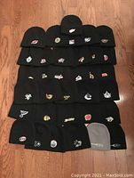 Photo showing complete set of 34 knit beanie hats (32 NHL plus 2 extras). Mostly black with NHL embroidered logos, two gray hats with Bud Light logos.