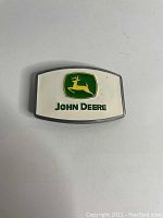 Front view of metal John Deere belt buckle showing green and yellow deer logo with 'JOHN DEERE' text on silver background.