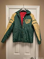 Front view of a green and tan men's hockey jacket with a red quilted lining and button closure, hanging on a door.