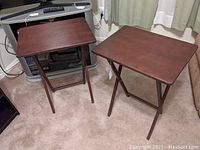Set of two dark stained folding TV tables standing on carpeted floor, showing rectangular tops and folding legs, in very good condition.