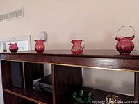 Four cranberry glass pieces displayed on top of a wooden shelf to show overall set