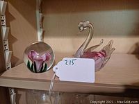 Photo showing spherical art glass paperweight with pink calla lilies and the pink glass swan figurine on a wood shelf.