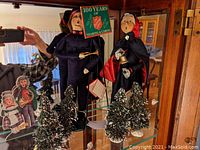 Two Salvation Army figurines standing behind four small flocked Christmas trees; male holding green sign, female in cape.