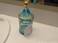 Blue enameled glass jar with floral design and gold accents, with lid topped by gold knob, shown from front.