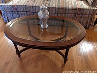 Front view of the oval coffee table with glass top and wooden legs, showing the beveled edge and cross braces underneath.