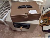 Brown steel cash box closed with black handle and latch, placed on top of white fireproof box