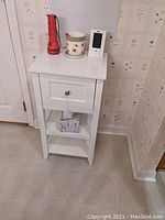 Photo showing the white wooden one drawer stand with a knob on the drawer, two open shelves, and items on top (not included)