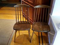 Two antique wooden chairs with spindle backs and contoured seats positioned side by side on a hardwood floor.