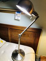 Full view of the articulating lamp showing the arm, base and lighted dome-shaped shade on a bed.