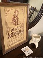 Framed vintage advertising sign for Dent's Toothache Gum alongside a ceramic tooth-shaped pencil holder, displayed on wooden furniture with a mirror in background.
