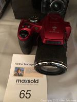 Red Fujifilm FinePix S9950W digital camera angled from front, showing lens and grip handle with maxsold lot number card.