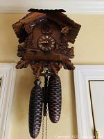 Front view of German wooden cuckoo clock mounted on wall, showing carved details and pinecone weights.