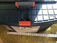 Closed toolbox showing black plastic with orange latch and measuring tape beside for size reference (approx 20 inches wide).