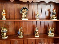 Front view of all eight Hummel figurines arranged on two wooden shelves against a paneled wall.