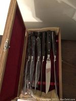 Set of six steak knives stored inside a wooden box with red velvet lining, showing knives mostly wrapped in plastic.