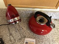 Red KitchenAid hand mixer with two beaters attached and an extra beater beside it, next to a red metal tea kettle with a black handle.