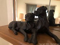 Photo of the large black Mastiff dog statue showing two dogs lying side by side from the front angle on a wooden surface with a mirror behind.