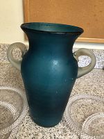 Teal glass ginger jar with two frosted handles surrounded by clear beaded luncheon plates on countertop.