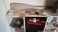 Wide angle photo showing the wooden paddle resting on top of shelving with various assorted game booklets and cards displayed below.