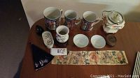 All items displayed together: three mugs, creamer, tea bag holder, bowls, canister, wooden piece, fan and silk panel
