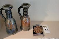 Two Suzy Atkins clay water pitchers side by side on white surface with a small round item next to them for scale.