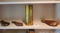 Four items displayed on a shelf: three powder horns and one large brass spent shell casing. Each powder horn varies in material with two metallic and one leather horn.