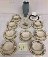 Photo showing the tall blue vase, 7 floral tea cups with matching saucers, one black and white tea cup with saucer, and one small decorative plate arranged on a white surface.