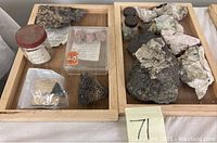 Top-down photo showing two wooden trays. Left tray contains various mineral samples including small jars, rock fragments and pink mineral pieces in plastic container with handwritten label. Right tray holds larger chunks of white and gray mineral rocks.
